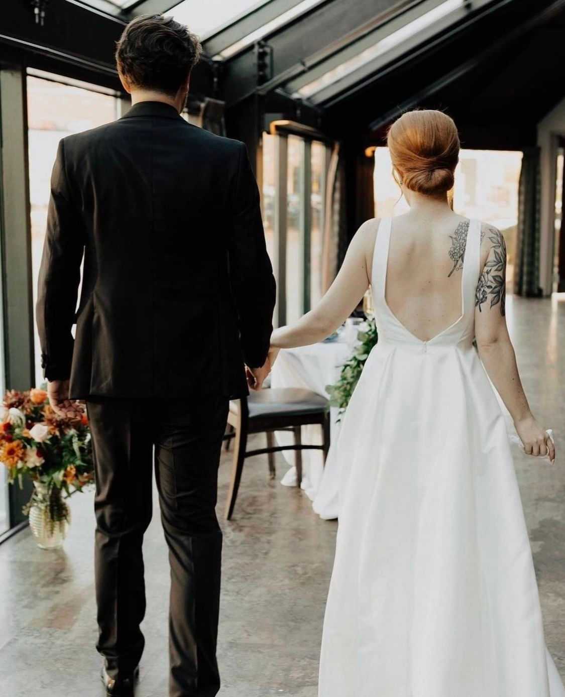 Couple in elegant wedding attire holding hands in a bright, glass-walled venue with floral decor.