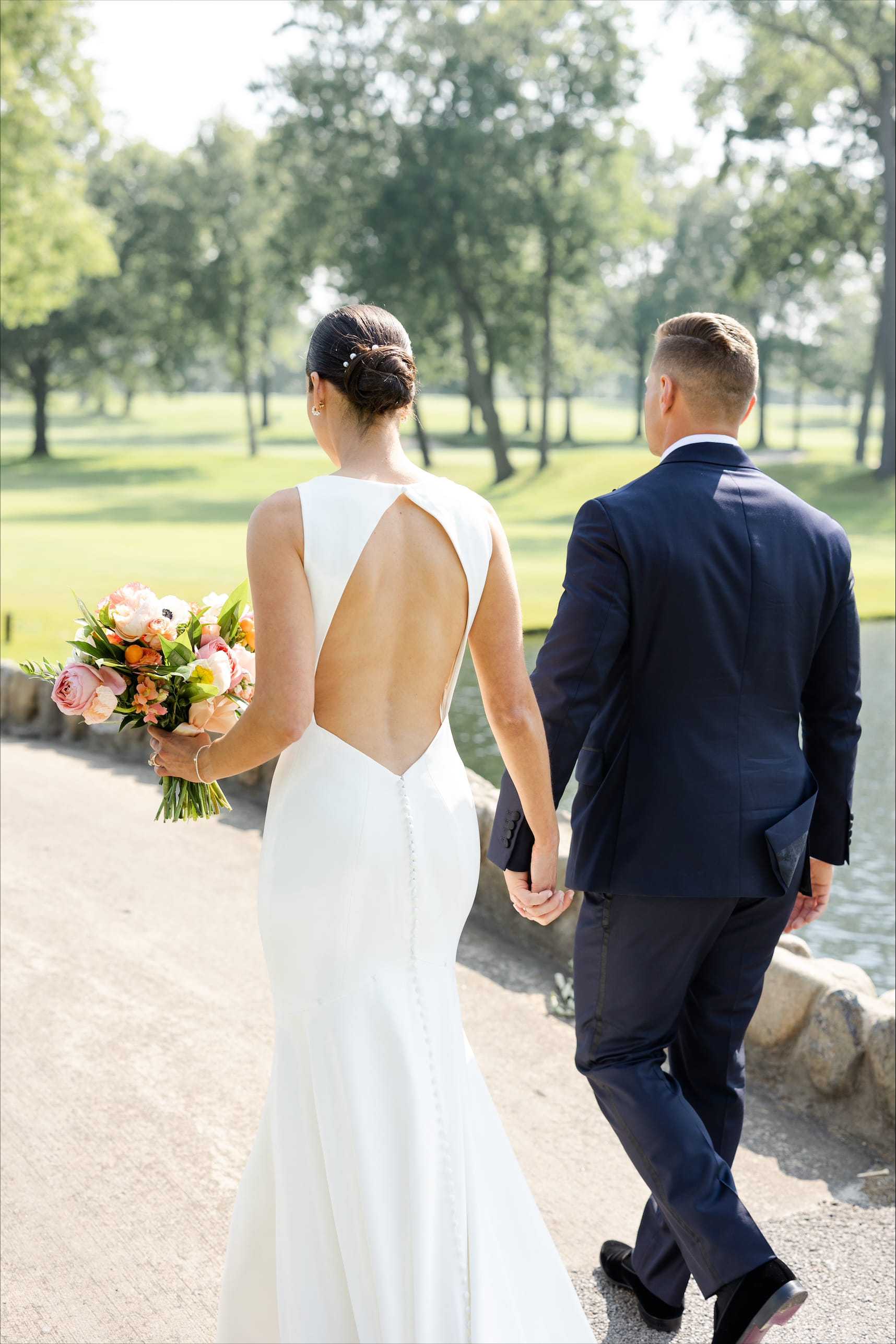 Bride and groom walking hand in hand near a pond, bride holding a bouquet.